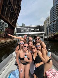Group of smiling friends in swimsuits on a pontoon boat cruising a downtown Fort Lauderdale canal under a raised drawbridge with high-rise buildings along the waterway
