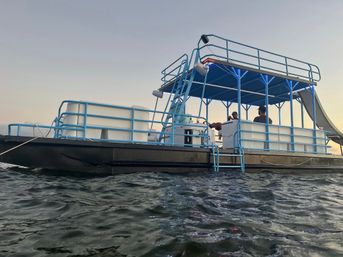 Double-deck pontoon boat with bright blue railings and slide floating on choppy lake water at sunset, passengers relaxing on the open upper and lower decks