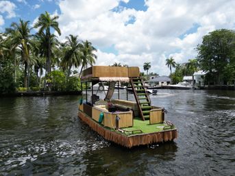 Tiki-style pontoon boat with grassy deck and thatch trim cruising a palm-lined Florida canal under a partly cloudy sky