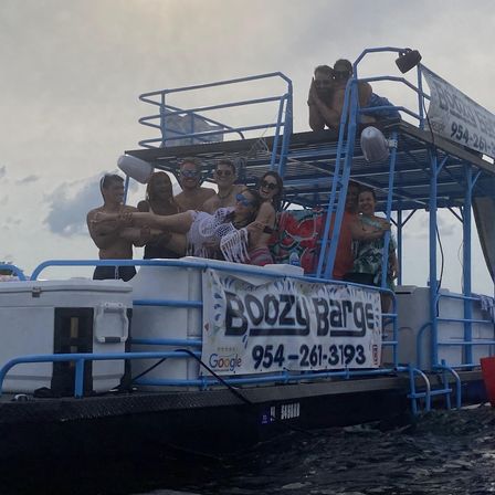 Group of friends partying on a blue two-level pontoon party boat in South Florida, smiling and posing as they hold a woman horizontally on deck over open water under a cloudy sky.