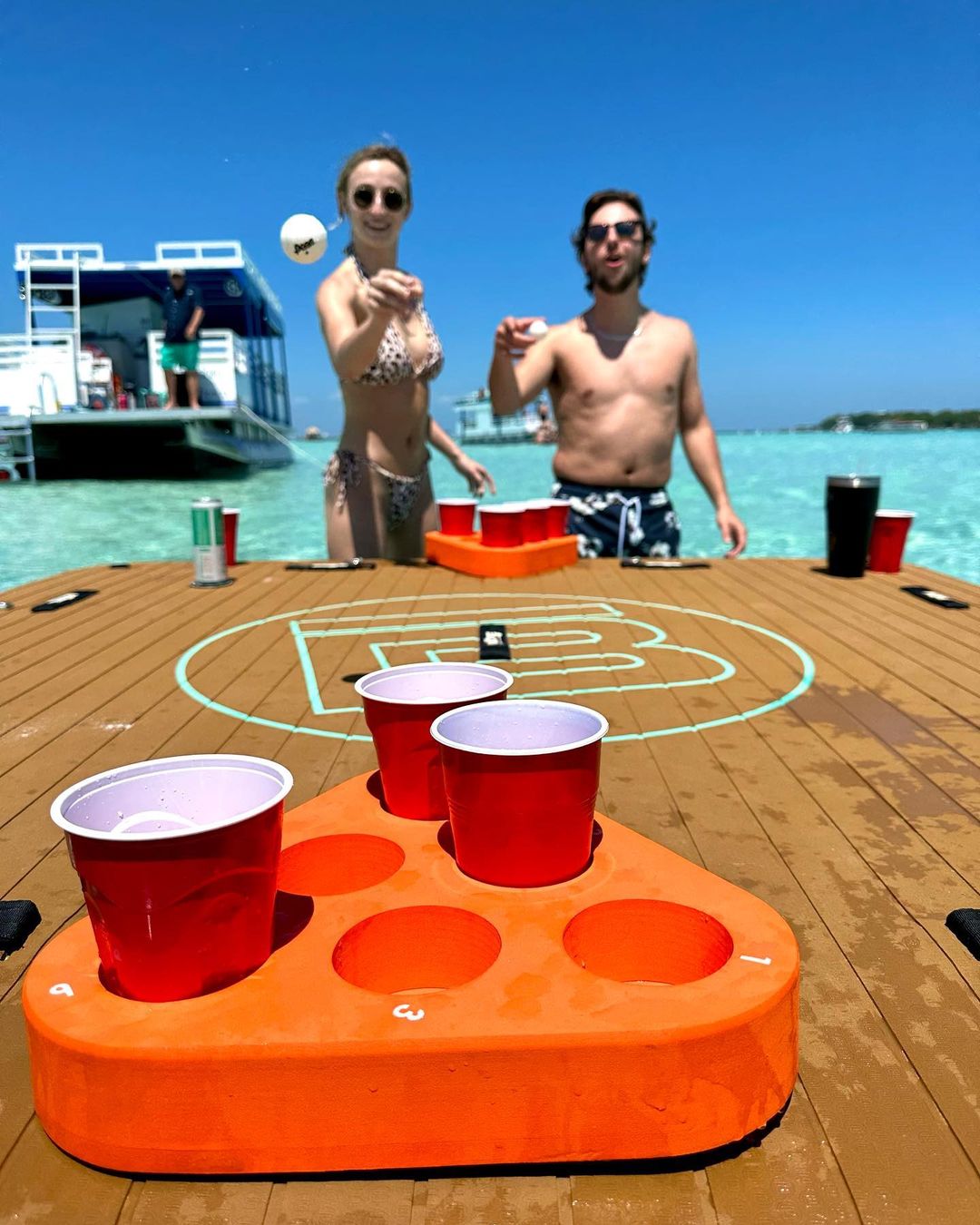 Two people playing beer pong with red cups on an orange floating rack on a wooden game table in clear turquoise water, boat and sunny blue sky in the background