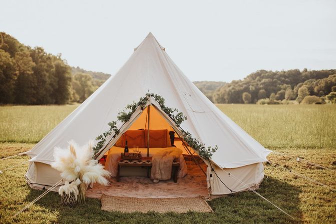 Inviting white bell tent glamping setup in a sunlit meadow — boho interior with a cozy bed, faux fur throw, bottle of wine, pampas grass and eucalyptus garland, rolling wooded hills in the background