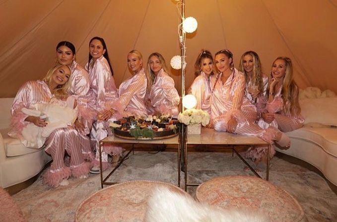 Group of women in matching pink satin striped feather-trim pajamas lounging in a cozy tent lounge around a low table with desserts, soft globe string lights, floral centerpiece and plush poufs — glamorous indoor pajama party.
