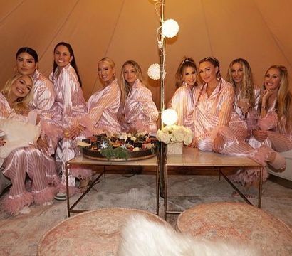 Group of women in matching pink satin striped feather-trim pajamas lounging in a cozy tent lounge around a low table with desserts, soft globe string lights, floral centerpiece and plush poufs — glamorous indoor pajama party.