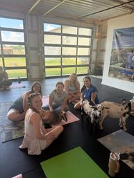 Group of smiling women on yoga mats in a bright barn-style studio enjoying a goat yoga session, petting a brown goat on a mat while two spotted goats wander nearby with large garage doors opening to a grassy farm outside.