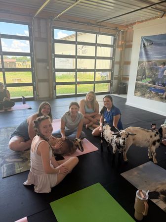 Group of smiling women on yoga mats in a bright barn-style studio enjoying a goat yoga session, petting a brown goat on a mat while two spotted goats wander nearby with large garage doors opening to a grassy farm outside.
