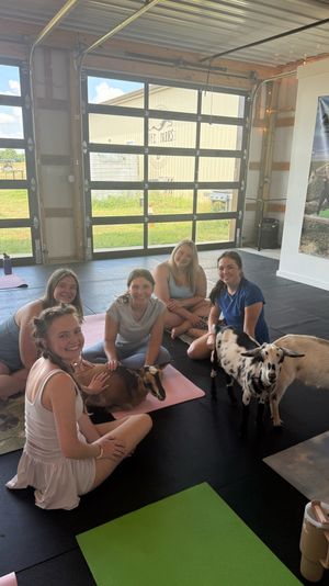 Group of smiling women on yoga mats in a bright barn-style studio enjoying a goat yoga session, petting a brown goat on a mat while two spotted goats wander nearby with large garage doors opening to a grassy farm outside.