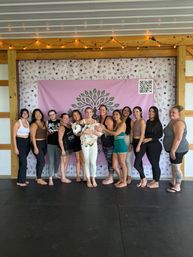 Barefoot group of women in yoga attire in a covered wooden pavilion doing goat yoga, holding a small goat in front of a pink tree-logo backdrop under string lights.