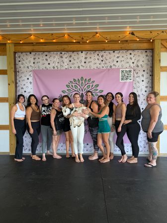 Barefoot group of women in yoga attire in a covered wooden pavilion doing goat yoga, holding a small goat in front of a pink tree-logo backdrop under string lights.