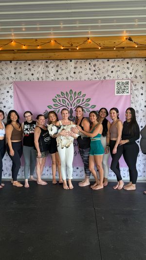 Barefoot group of women in yoga attire in a covered wooden pavilion doing goat yoga, holding a small goat in front of a pink tree-logo backdrop under string lights.