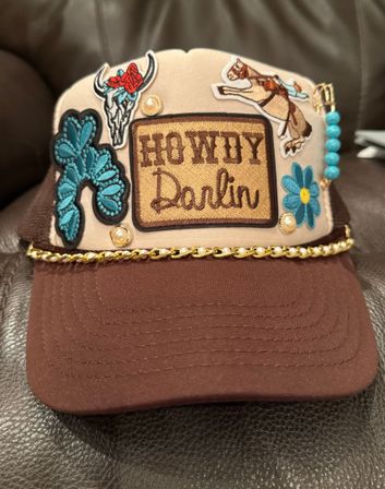 Brown-and-beige Western-style trucker hat with an embroidered "Howdy Darlin" patch, turquoise appliqué flowers, steer skull and horse patches, turquoise bead dangles and gold chain trim — cowgirl fashion accessory.
