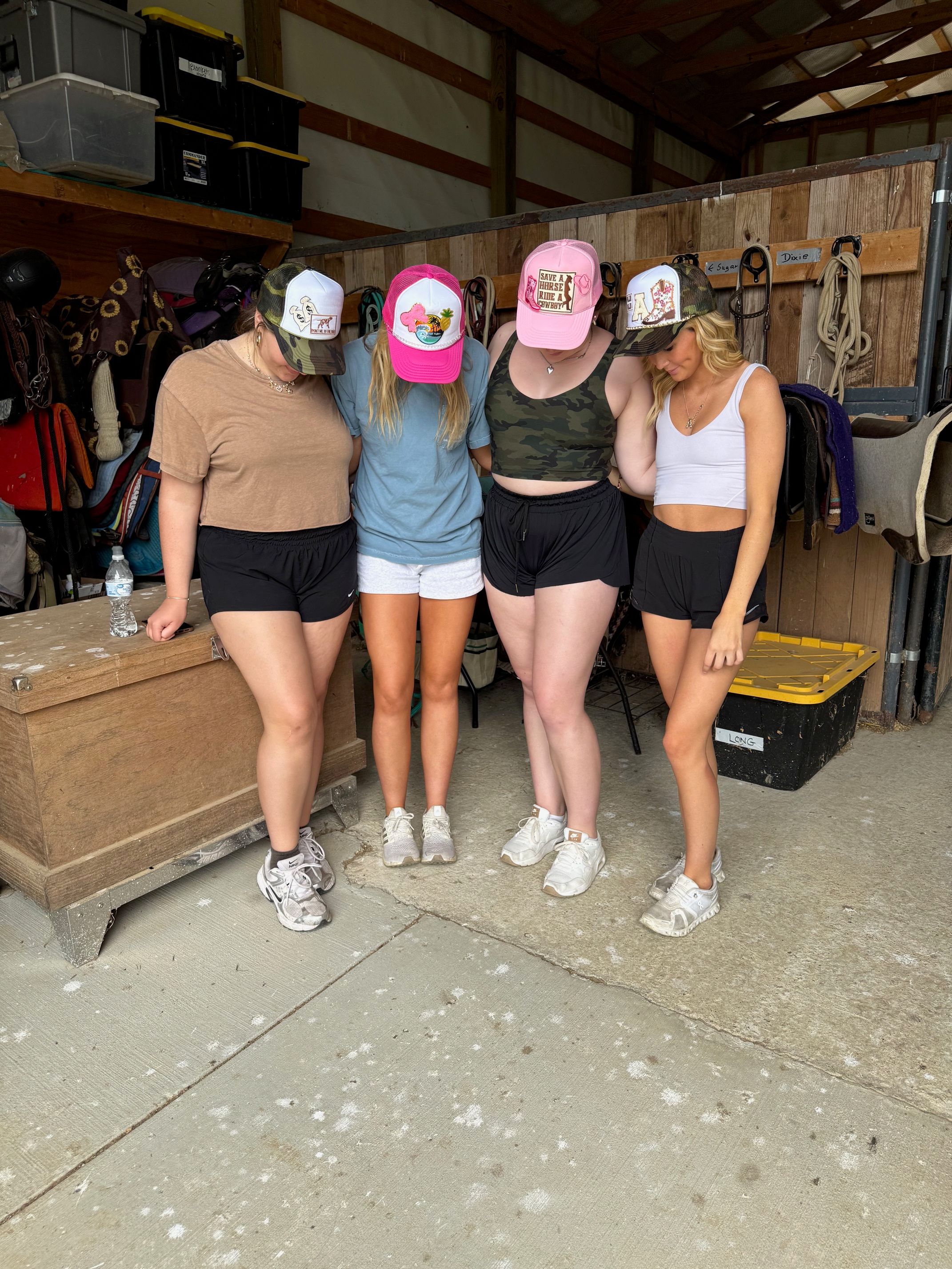 Four friends in a horse barn wearing colorful trucker hats and summer athletic shorts, standing close with heads bowed in front of saddles, bridles, and tack.