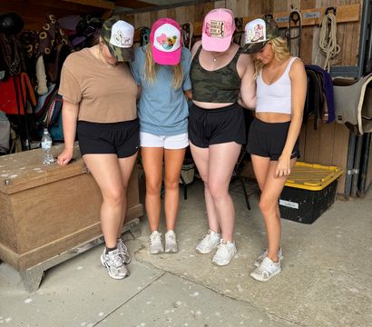Four friends in a horse barn wearing colorful trucker hats and summer athletic shorts, standing close with heads bowed in front of saddles, bridles, and tack.