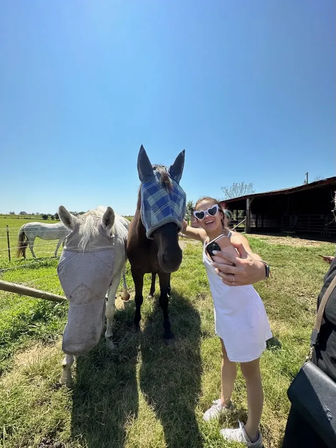 Person in a white sundress and heart-shaped sunglasses taking a selfie with two horses wearing protective fly masks in a sunny country pasture beside a barn