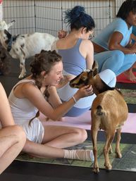 Smiling woman in white activewear kneeling on a yoga mat and gently petting a small brown goat during an indoor goat yoga class with other participants on mats.