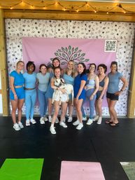 Smiling group of women in blue activewear posing at an indoor goat yoga class in front of a pink backdrop with a green tree logo and string lights, one woman holding a small white goat.