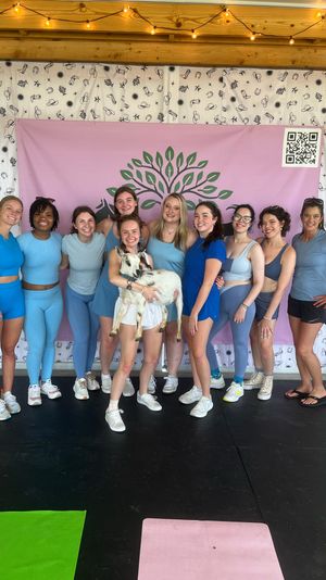 Smiling group of women in blue activewear posing at an indoor goat yoga class in front of a pink backdrop with a green tree logo and string lights, one woman holding a small white goat.