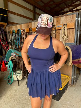 Woman in navy sundress wearing a decorated trucker cap reading Save a Horse Ride a Cowboy, standing in a horse barn tack room with saddles, bridles, ropes and horse blankets