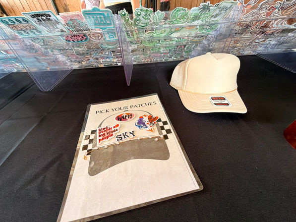 Craft market vendor table with clear acrylic rack of embroidered patches, a 'pick your patches' selection sheet and a beige OTTO trucker hat on a black tablecloth
