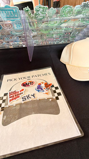 Craft market vendor table with clear acrylic rack of embroidered patches, a 'pick your patches' selection sheet and a beige OTTO trucker hat on a black tablecloth