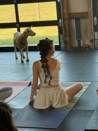 Person seated on a yoga mat in a rustic barn studio during goat yoga — a curious goat stands by an open garage door overlooking a sunny grassy pasture.