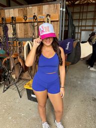 Smiling person in a bright blue athletic crop top and shorts tipping a pink-and-white trucker hat inside a rustic horse barn tack room with saddles, bridles and horse blankets.