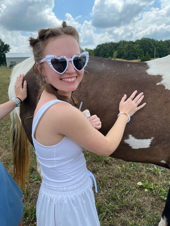 Smiling woman in a white dress wearing heart-shaped "Bride to Be" sunglasses, petting a brown-and-white horse in a sunny rural pasture under a partly cloudy sky.
