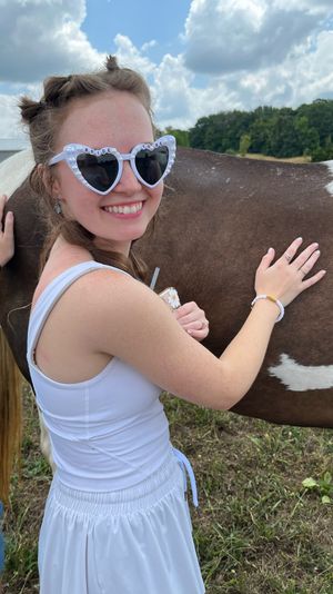Smiling woman in a white dress wearing heart-shaped "Bride to Be" sunglasses, petting a brown-and-white horse in a sunny rural pasture under a partly cloudy sky.