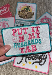 Hand holding a rectangular embroidered novelty patch reading PUT IT ON MY HUSBANDS TAB in pink and teal, surrounded by playful western- and cowgirl-themed patches on a carpeted surface.