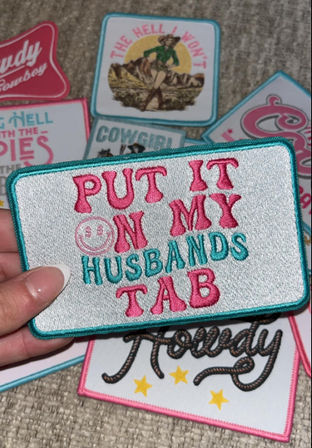 Hand holding a rectangular embroidered novelty patch reading PUT IT ON MY HUSBANDS TAB in pink and teal, surrounded by playful western- and cowgirl-themed patches on a carpeted surface.