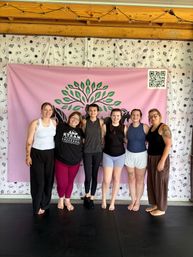Six women standing barefoot in a row smiling with arms around each other in a wellness studio, in front of a pink banner with a green tree logo and cowboy-patterned white curtain, wooden beam ceiling and string lights overhead.