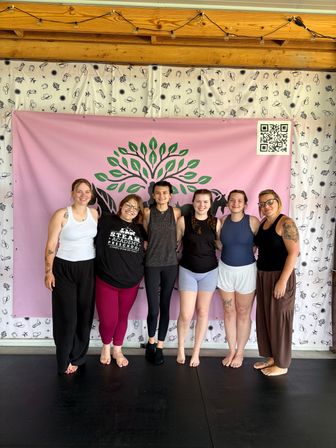 Six women standing barefoot in a row smiling with arms around each other in a wellness studio, in front of a pink banner with a green tree logo and cowboy-patterned white curtain, wooden beam ceiling and string lights overhead.