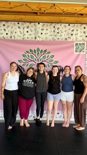 Six women standing barefoot in a row smiling with arms around each other in a wellness studio, in front of a pink banner with a green tree logo and cowboy-patterned white curtain, wooden beam ceiling and string lights overhead.