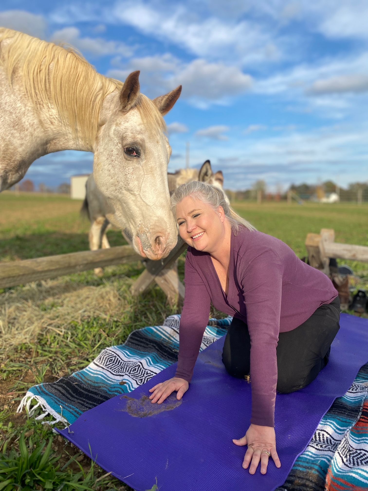 Smiling woman kneeling on a purple yoga mat on a colorful blanket in a sunny rural pasture as a light-colored horse gently nuzzles her over a wooden fence