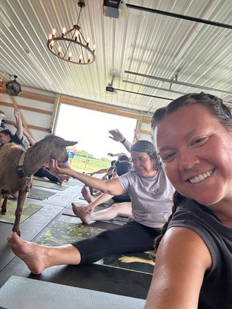 Smiling participant selfie at a goat yoga class in a bright open-sided barn — people on mats stretching as a curious goat nuzzles a hand under a metal-paneled ceiling and chandelier.
