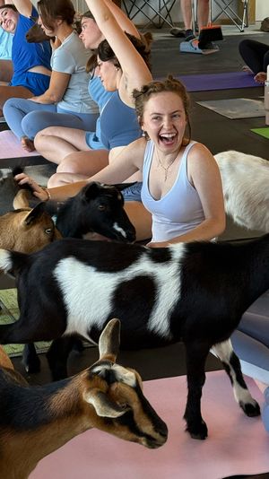 Smiling woman laughing and petting small goats during a lively indoor goat yoga class on colorful yoga mats