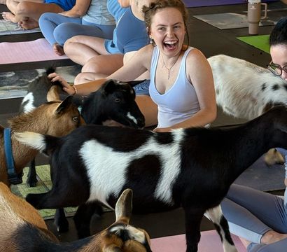 Smiling woman laughing and petting small goats during a lively indoor goat yoga class on colorful yoga mats