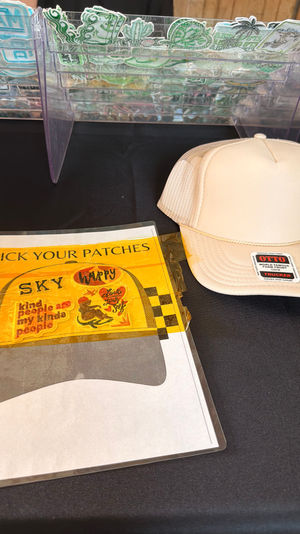 Vendor table with a beige trucker hat and a clear organizer holding colorful iron-on patches and a 'Pick Your Patches' sign.