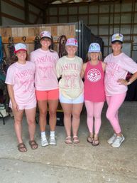 Five women posing in a barn-style stable, wearing pink Nashville T-shirts, colorful decorated trucker caps and casual shorts or leggings, smiling in front of horse tack and wooden stalls.