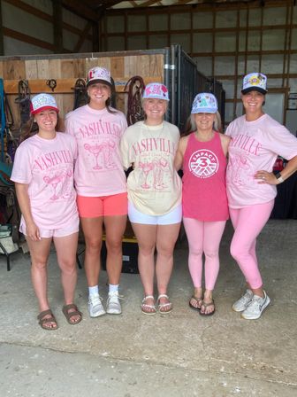Five women posing in a barn-style stable, wearing pink Nashville T-shirts, colorful decorated trucker caps and casual shorts or leggings, smiling in front of horse tack and wooden stalls.