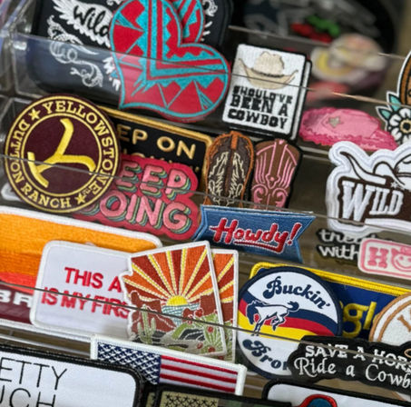 Bright, western-themed embroidered patches on a display rack — 'Howdy!' banner, bucking bronco, cactus sunset (Arizona-style), Yellowstone ranch emblem, American flag and other cowboy motifs