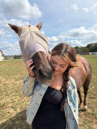 Person with braided hair in a denim jacket hugging a brown horse wearing a tan fly mask in a sunny rural pasture under a blue sky with fluffy clouds