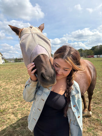 Person with braided hair in a denim jacket hugging a brown horse wearing a tan fly mask in a sunny rural pasture under a blue sky with fluffy clouds