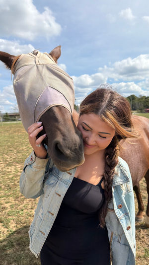 Person with braided hair in a denim jacket hugging a brown horse wearing a tan fly mask in a sunny rural pasture under a blue sky with fluffy clouds