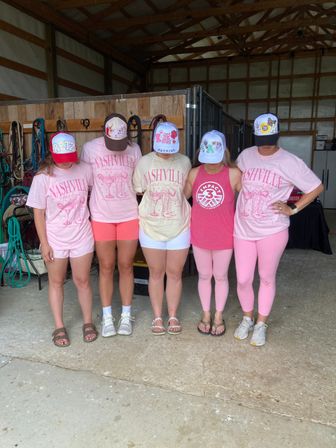 Five women wearing pink 'Nashville' tees and decorated trucker hats standing with heads down inside a wooden horse barn with bridles and tack on the wall.