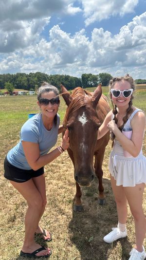 Two smiling women wearing sunglasses pet a chestnut horse with a white star in a sunny rural pasture under blue sky and puffy clouds, summer farm outing.