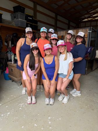 Eight smiling friends in colorful trucker hats pose inside a wooden horse barn beside saddles and tack, wearing casual summer athletic outfits.