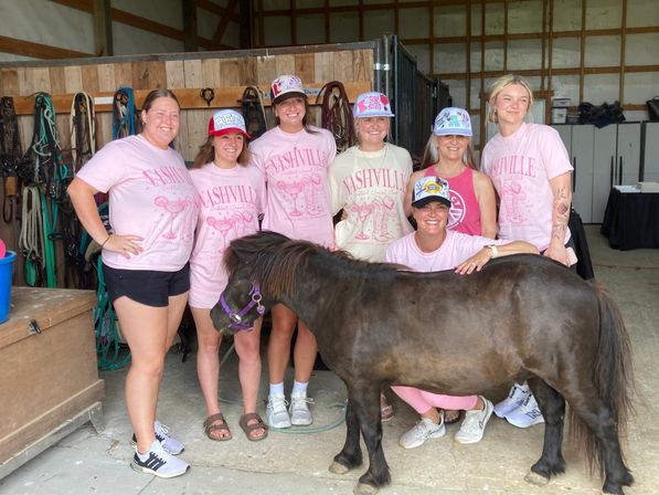 Smiling group of eight women in pink “Nashville” shirts and caps posing with a small black pony inside a wooden barn stable with bridles and tack on the wall.