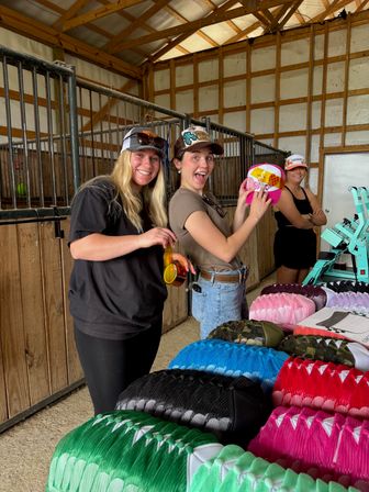 Three smiling women in a wooden barn browsing and holding colorful trucker caps stacked on a table next to a hat-press machine.