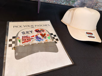 Beige trucker hat beside a "Pick Your Patches" display of colorful iron-on patches and a pearl chain on a black table — DIY hat-decorating station.